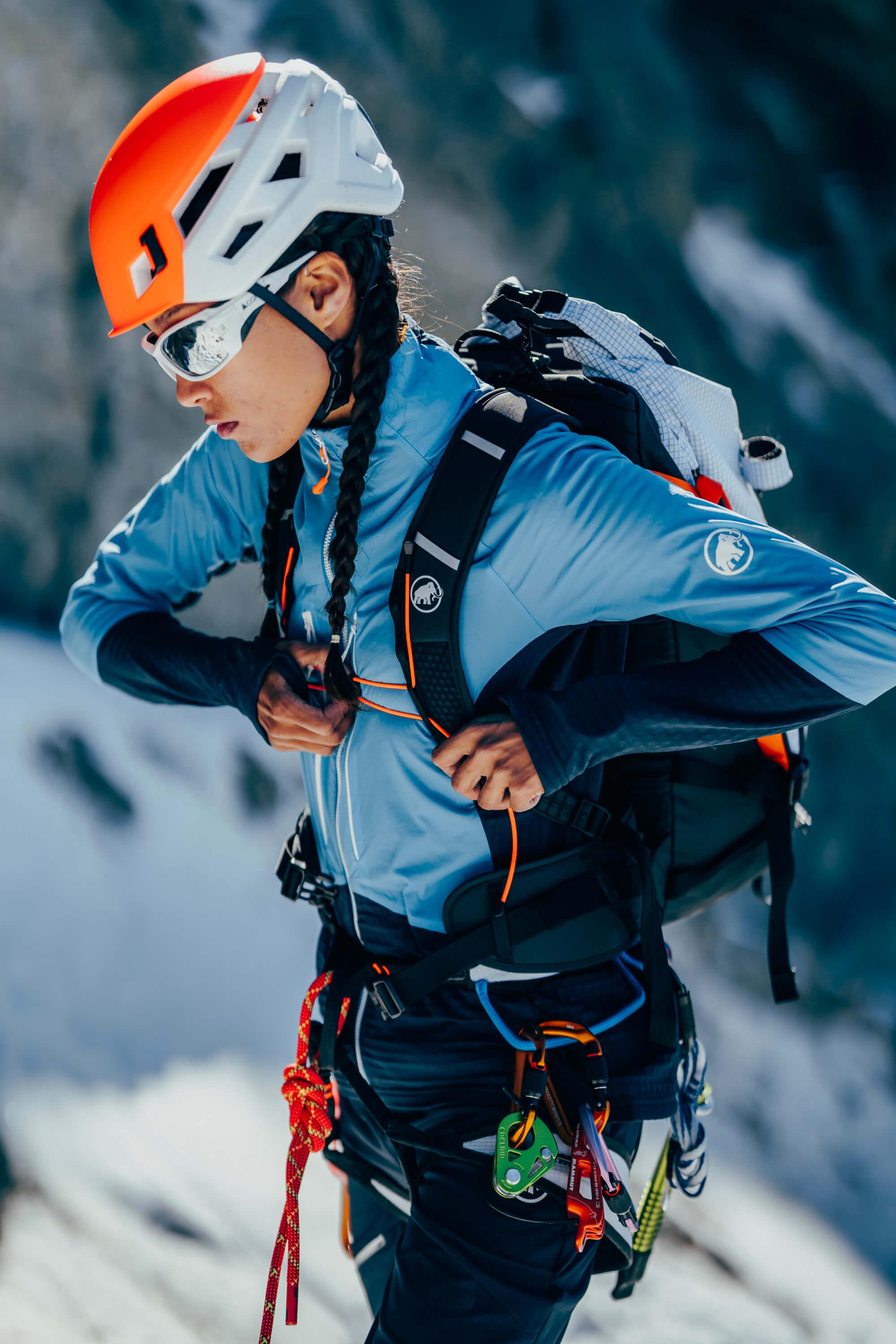 A Mammut climber wearing a helmet, sunglasses, and premium climbing gear adjusts harness straps on snowy mountain terrain.