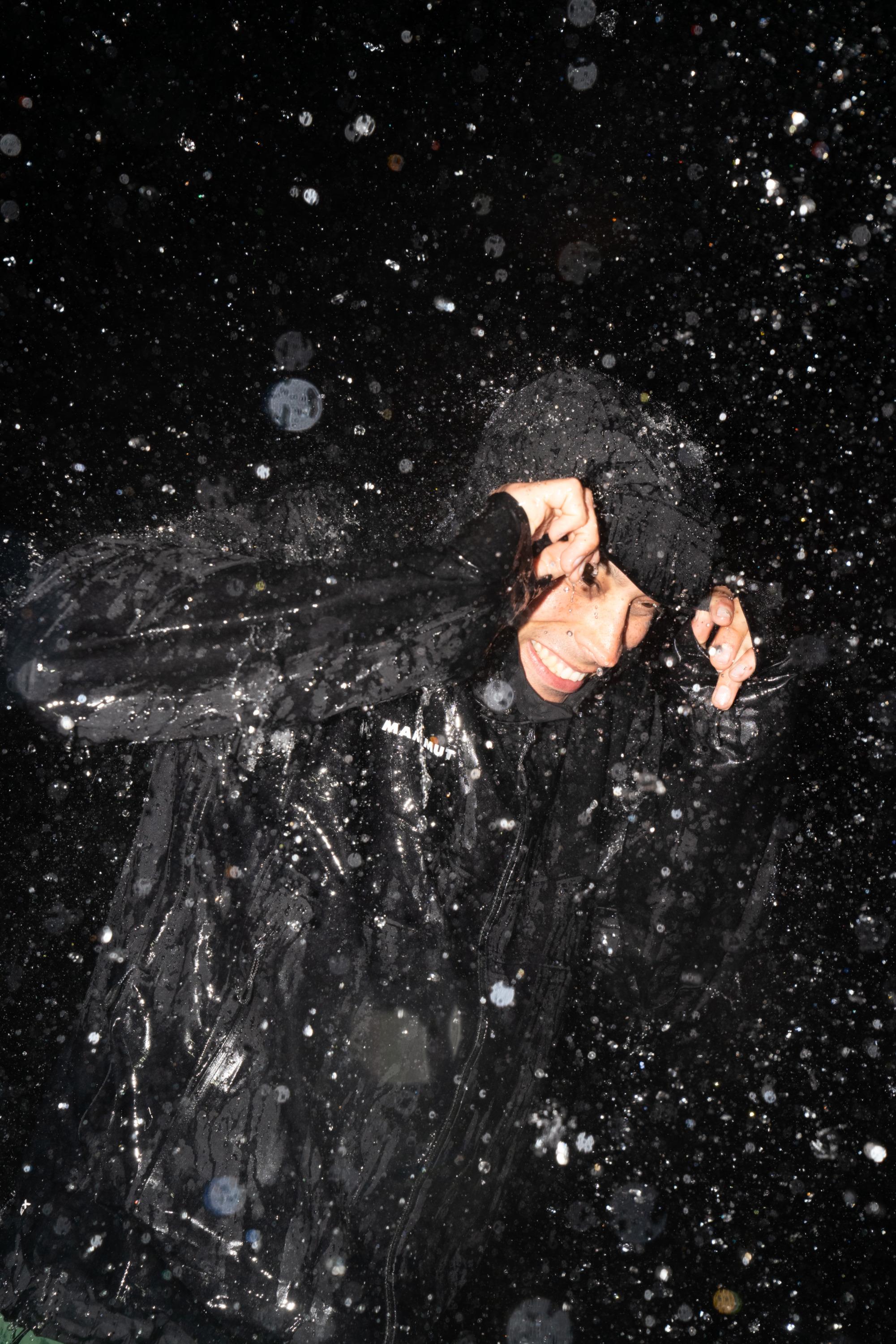 Person in a black Mammut raincoat, smiling and shielding their face while being splashed with water droplets at night.