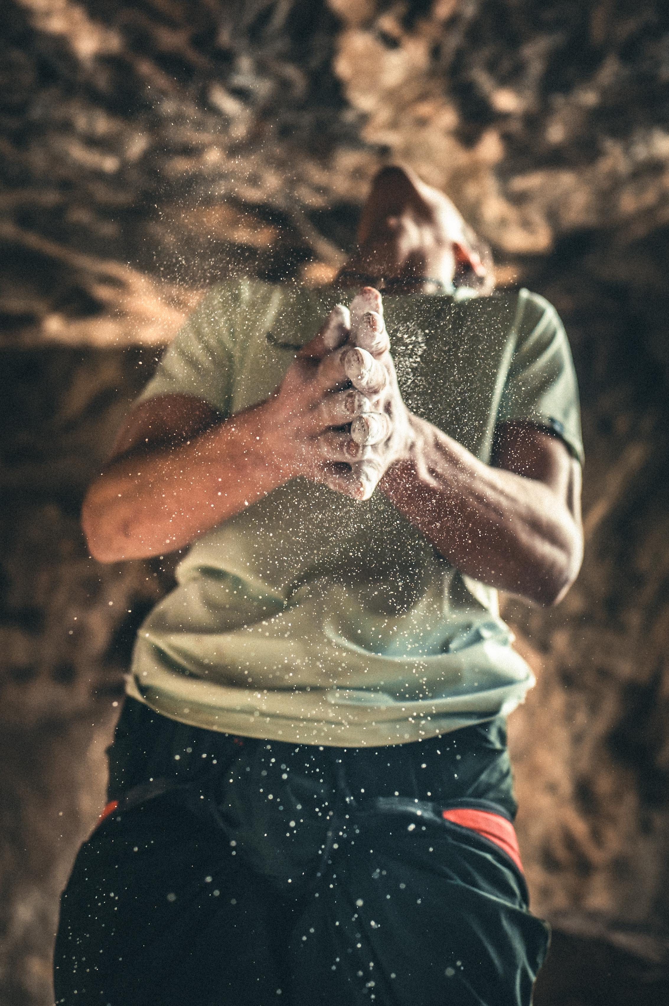 Person wearing Mammut climbing gear claps powdered hands in a cave, viewed from below. Dust particles are highlighted by light in the background.
