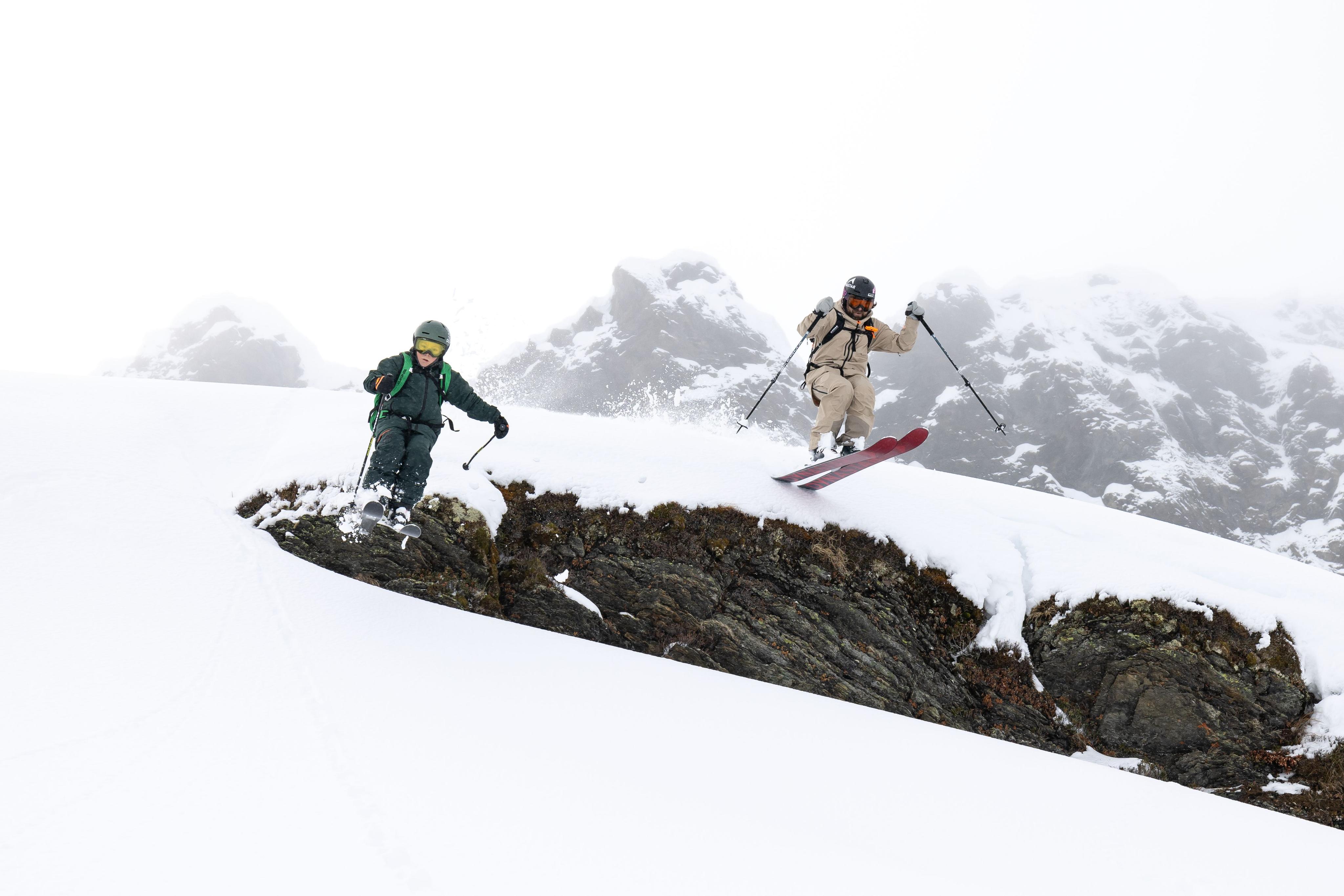 Whisky and his son, two skiers in vibrant Mammut gear navigate a snowy mountain with rocky patches, beneath a cloudy sky.