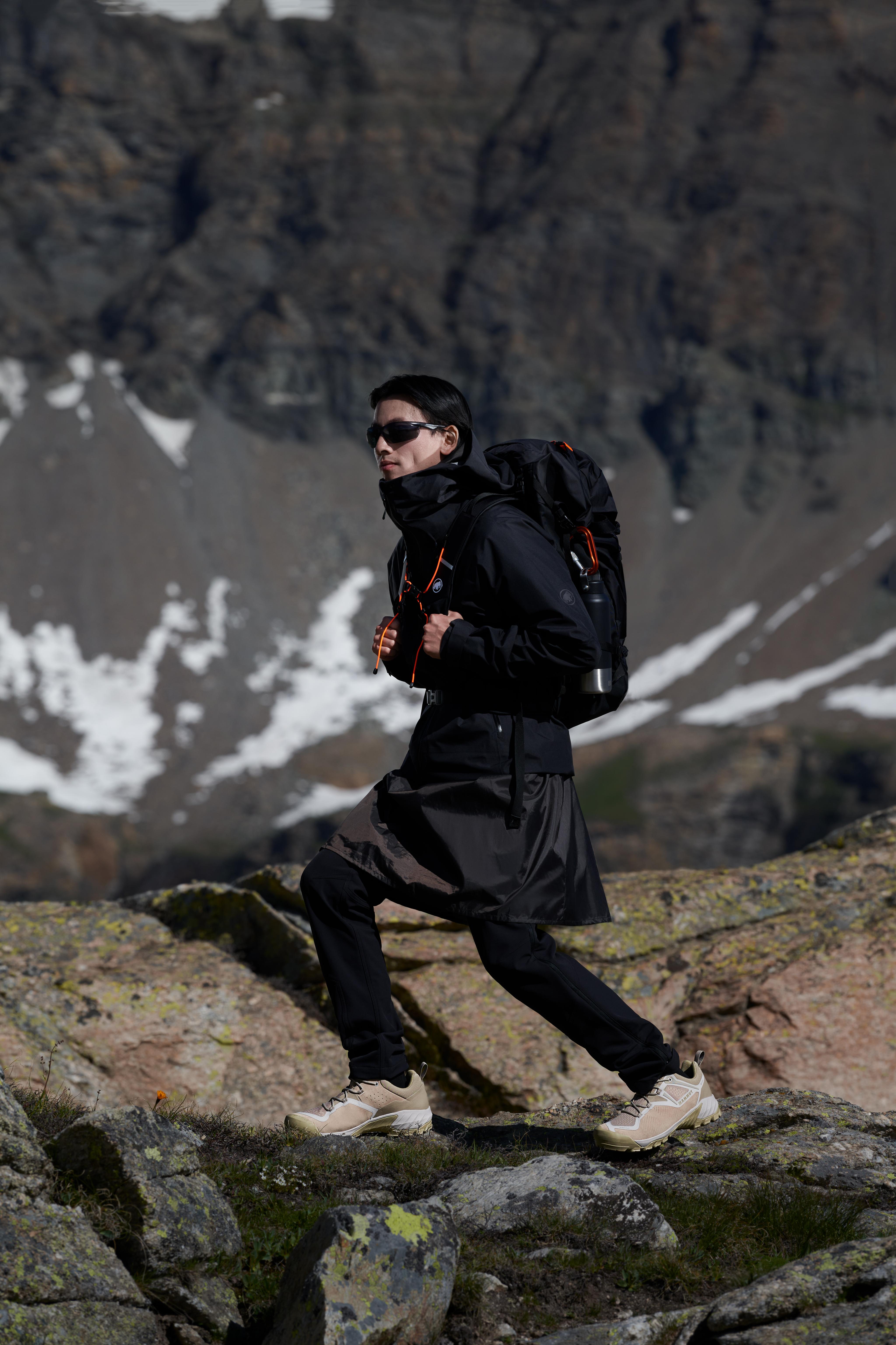 Person in Mammut black hiking gear with a backpack navigating rocky terrain, with snow-capped mountains in the background.