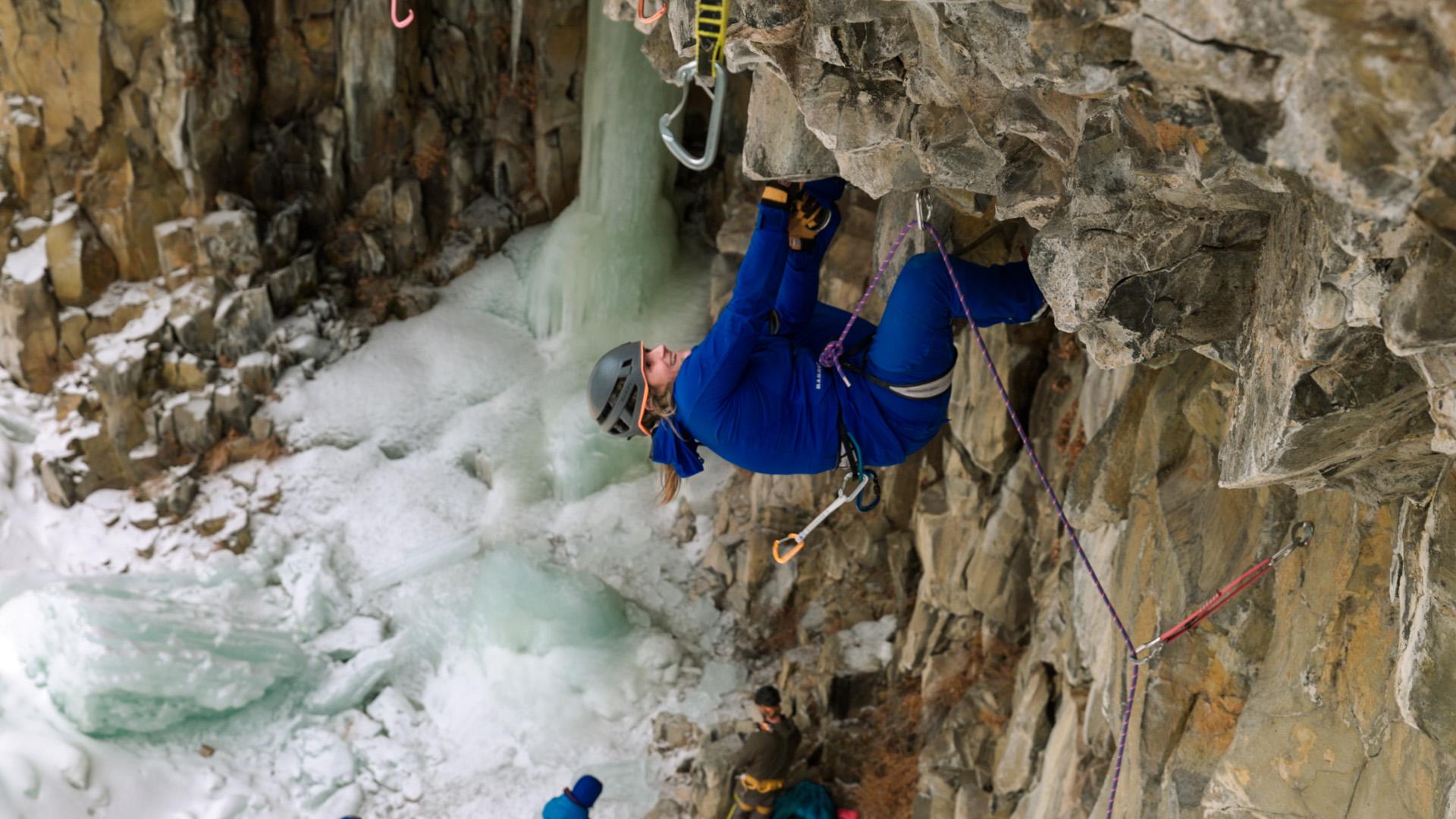Climber in blue Mammut gear ascends icy, vertical rock wall with ropes; snow and ice below, onlookers watching.