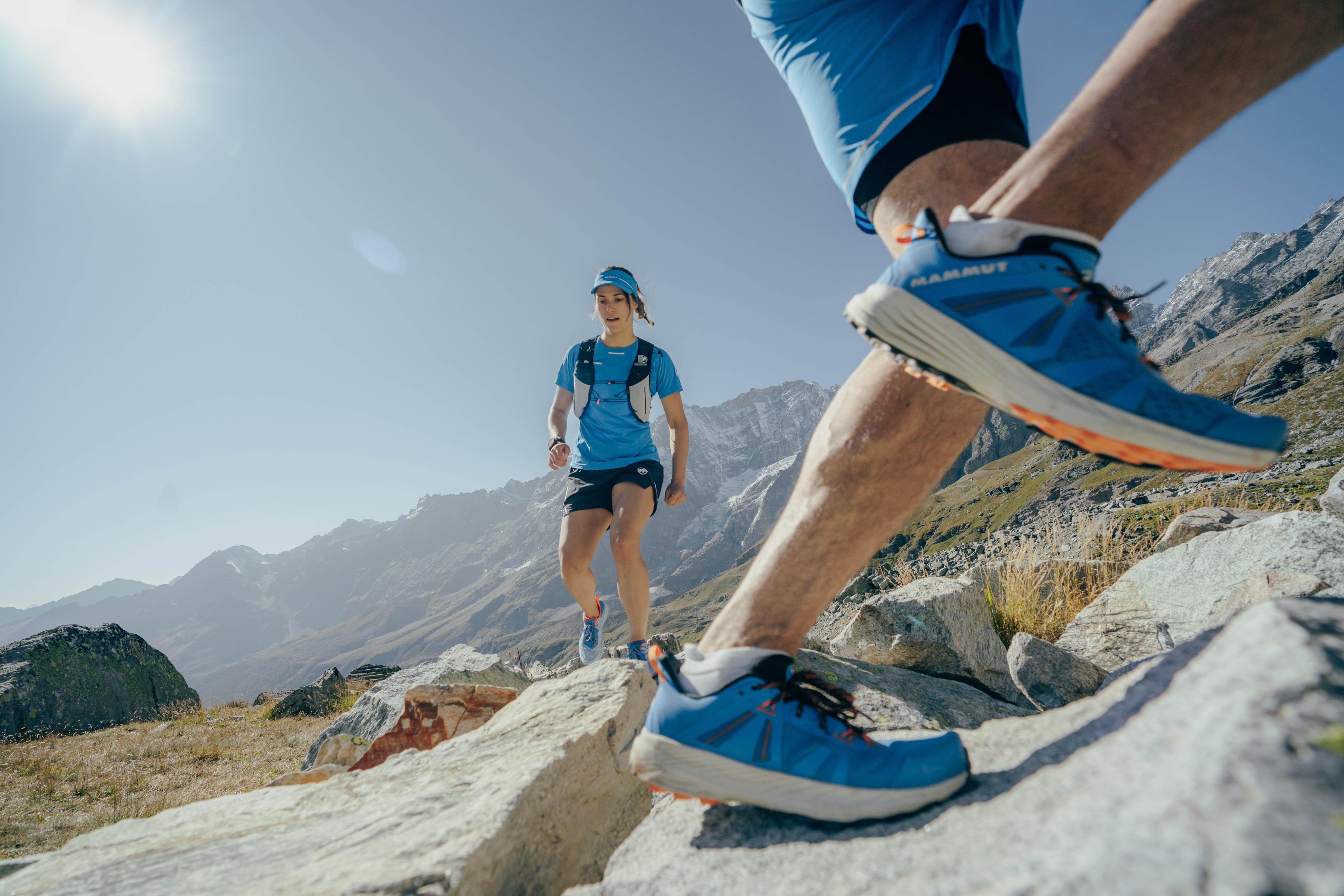 Two trail runners wearing blue Mammut gear navigating rocky terrain, with majestic mountains and a clear sky in the background.