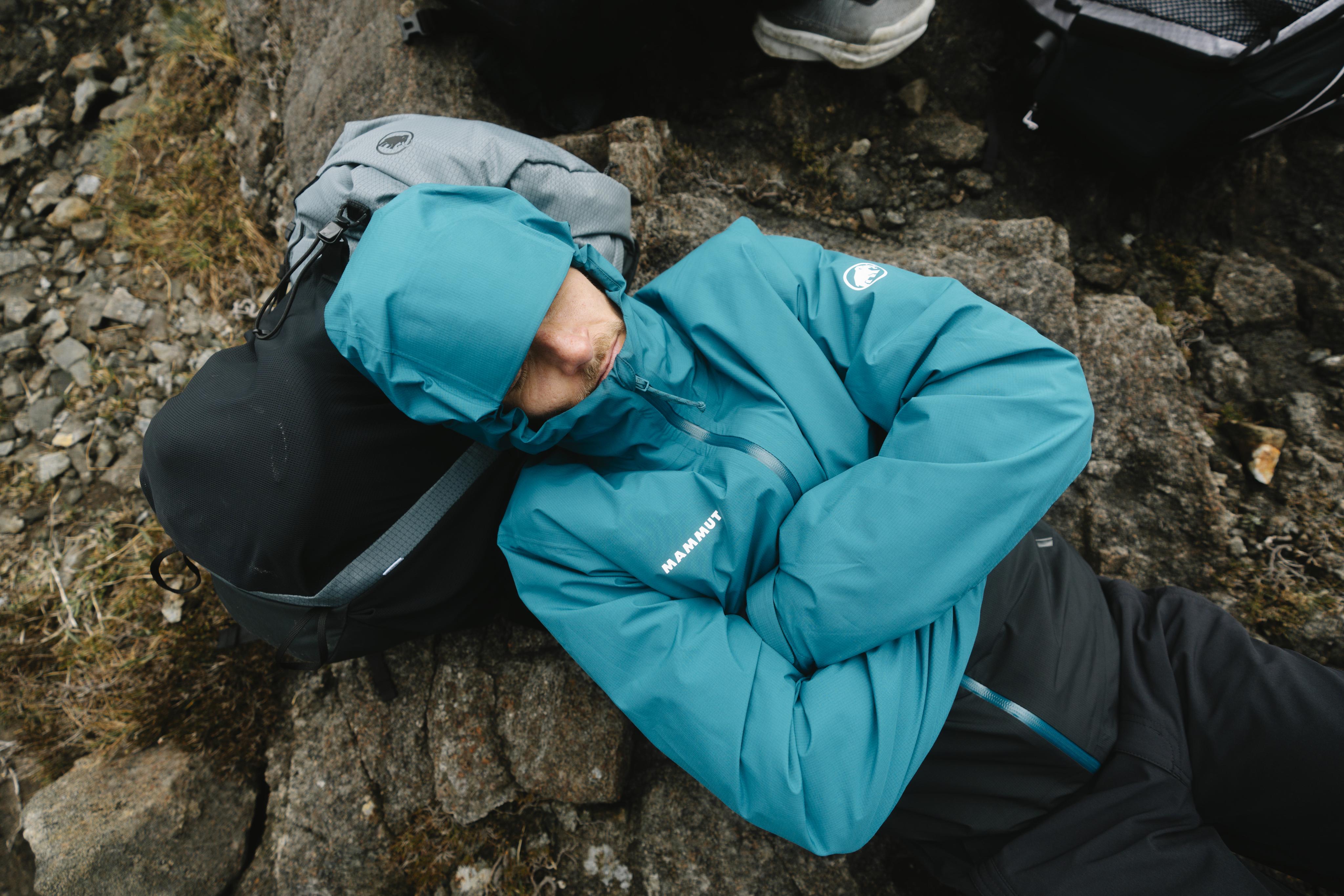 Person in a teal Mammut jacket with hood sleeping on rocky terrain outdoors, using a Mammut backpack as a pillow.