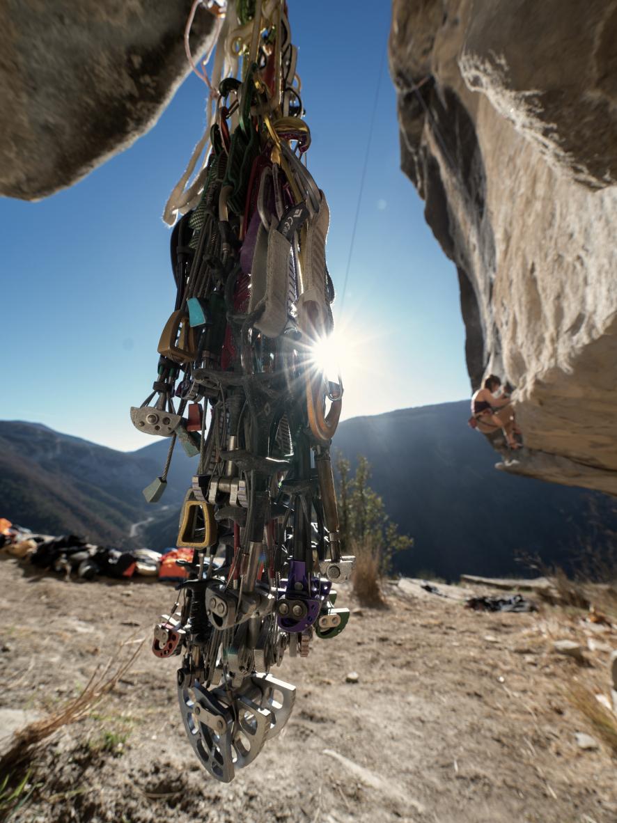 Mammut climbing gear hangs as a climber ascends a rugged rock face, with a bright sunlit valley backdrop.