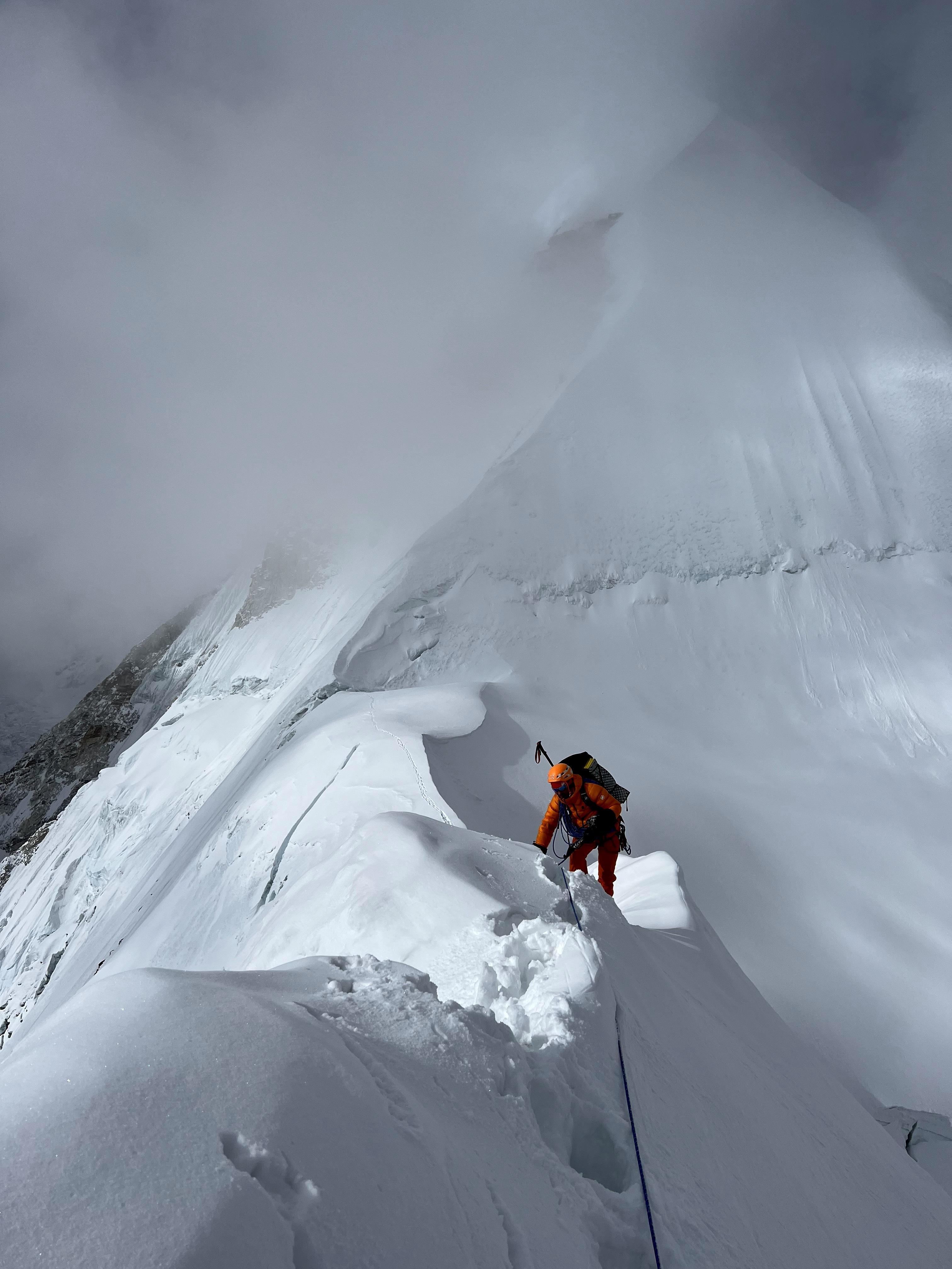 Mammut-sponsored climber in orange gear ascending a snowy, steep mountain ridge beneath cloudy skies.