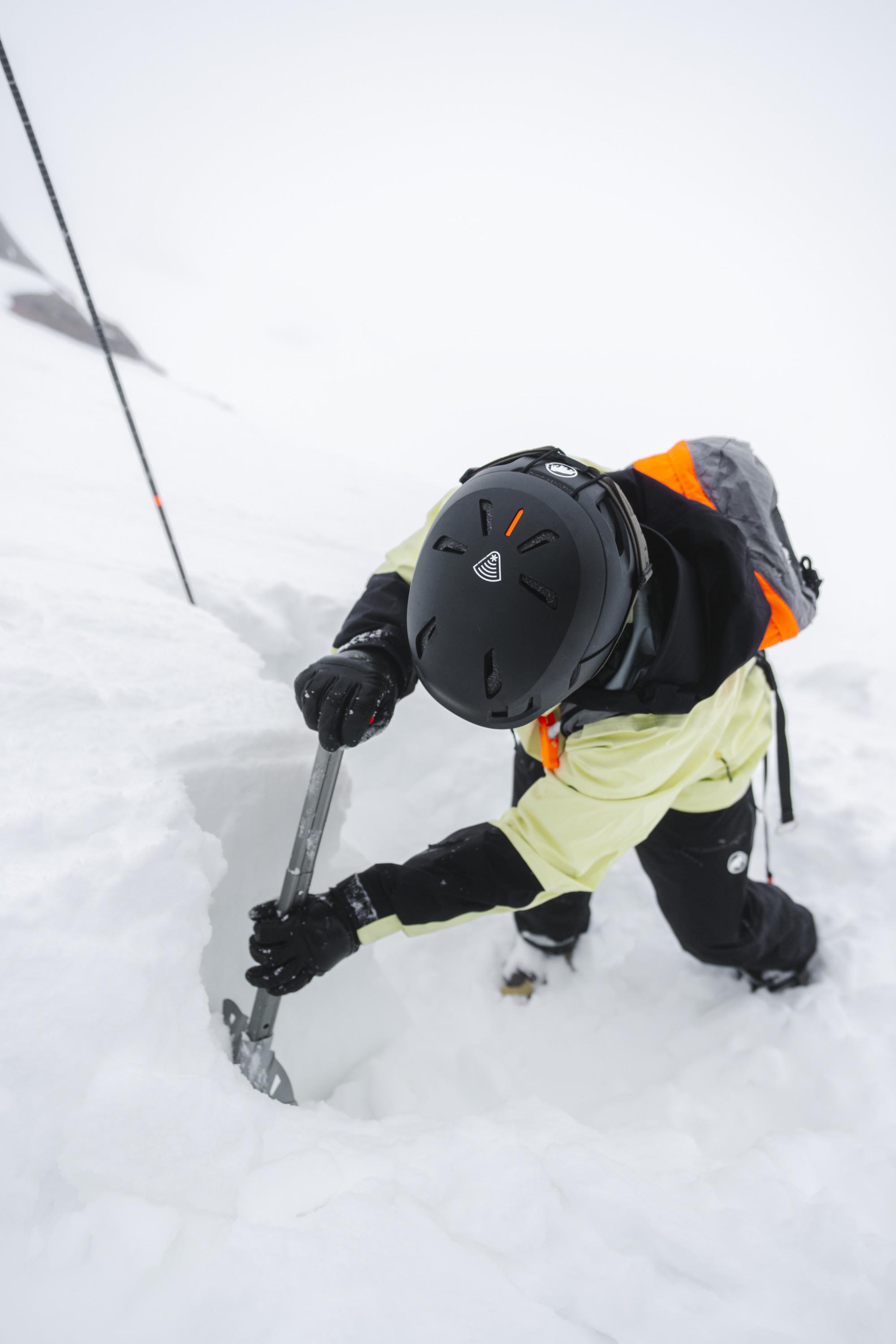 Gloved hand with a Mammut logo holding an orange snow shovel, efficiently digging into deep alpine snow.