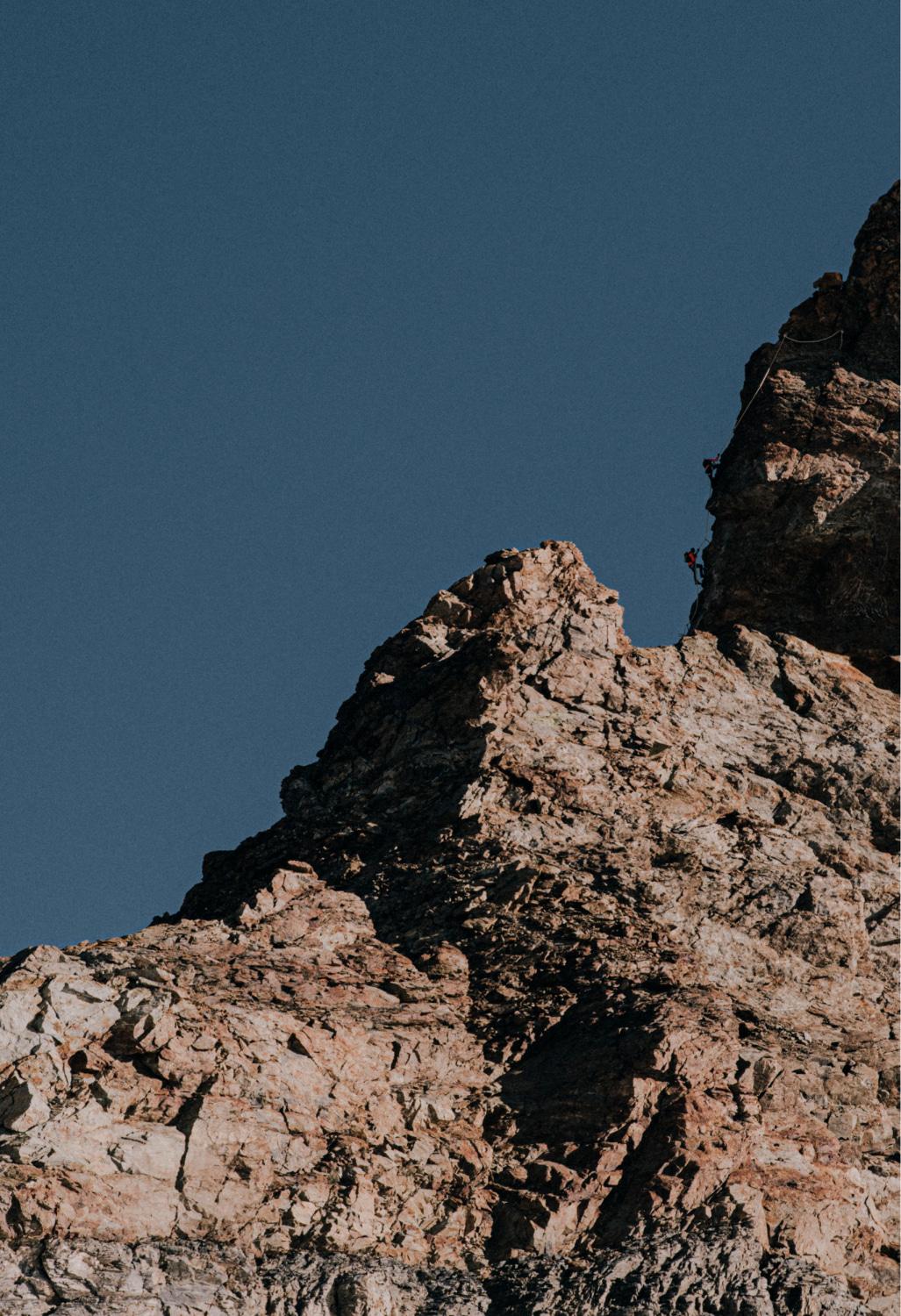 A lone climber wearing Mammut gear ascends a rugged rocky cliff against a clear blue sky.