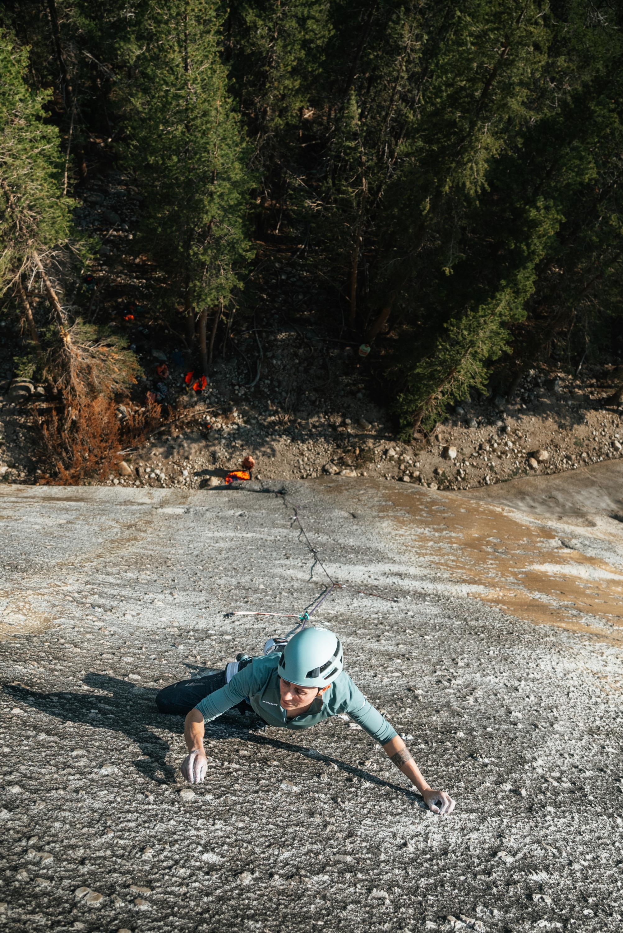 A climber wearing a Mammut helmet and gear ascends a steep, rocky cliff with a forested landscape in the background.