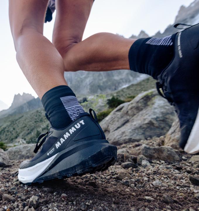 Close-up of a trail runner ascending a rocky incline, wearing black Mammut performance running shoes for mountain terrain.