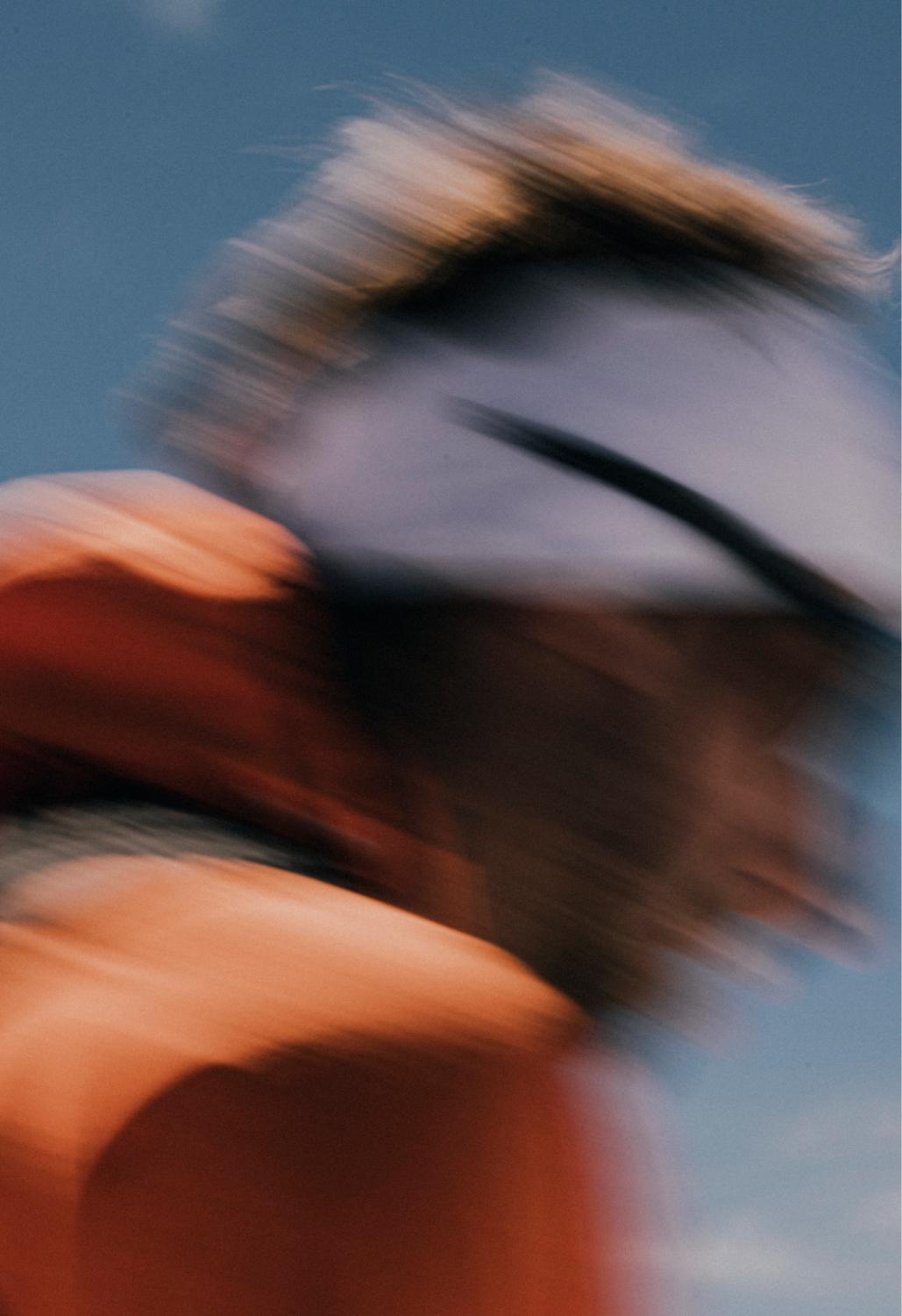 A dynamic, blurry image of a person in motion under a clear blue sky, wearing an orange Mammut jacket and a light purple visor.