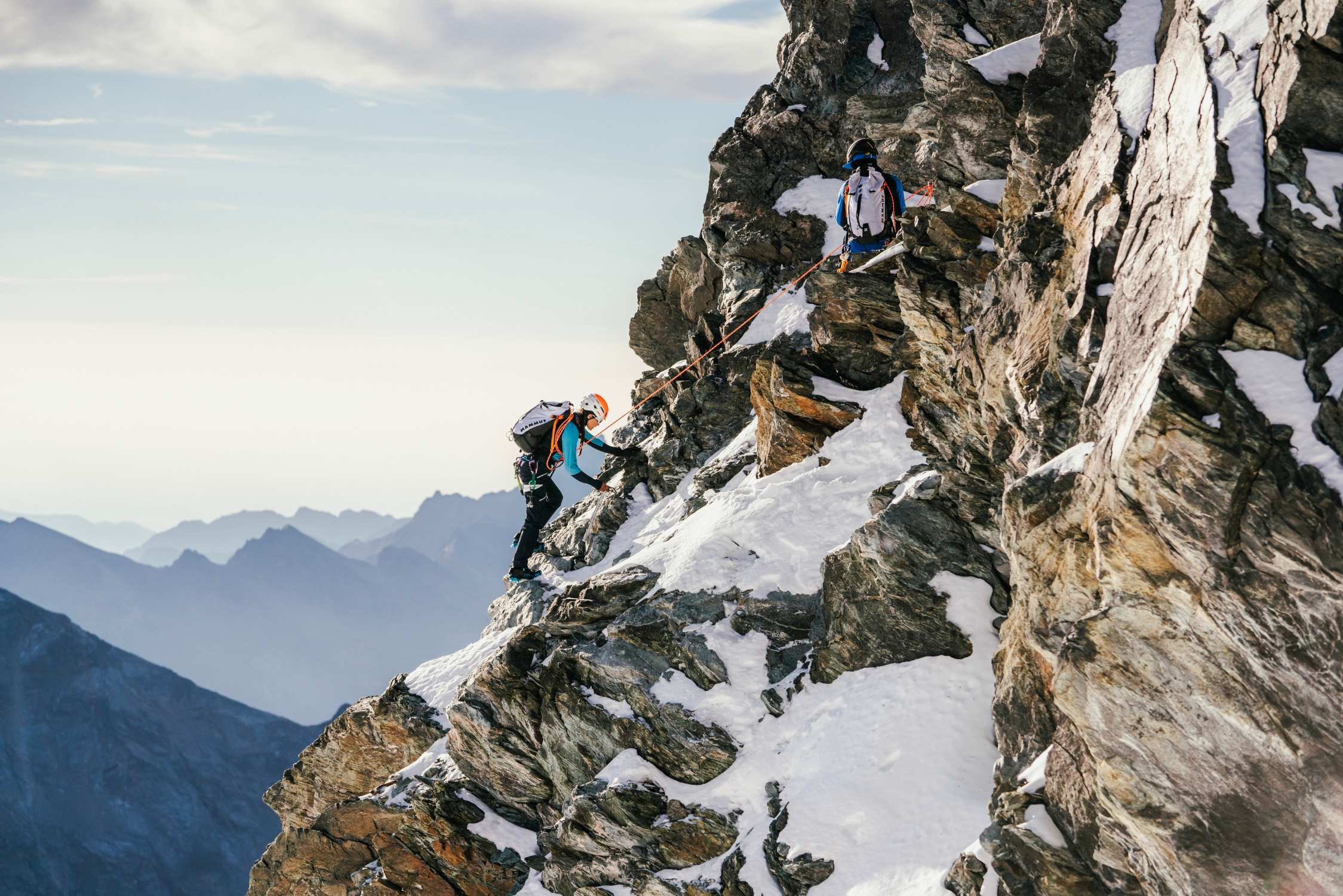 Two Mammut-clad climbers with gear ascend a snowy, rocky mountain slope, alpine peaks rising in the background.