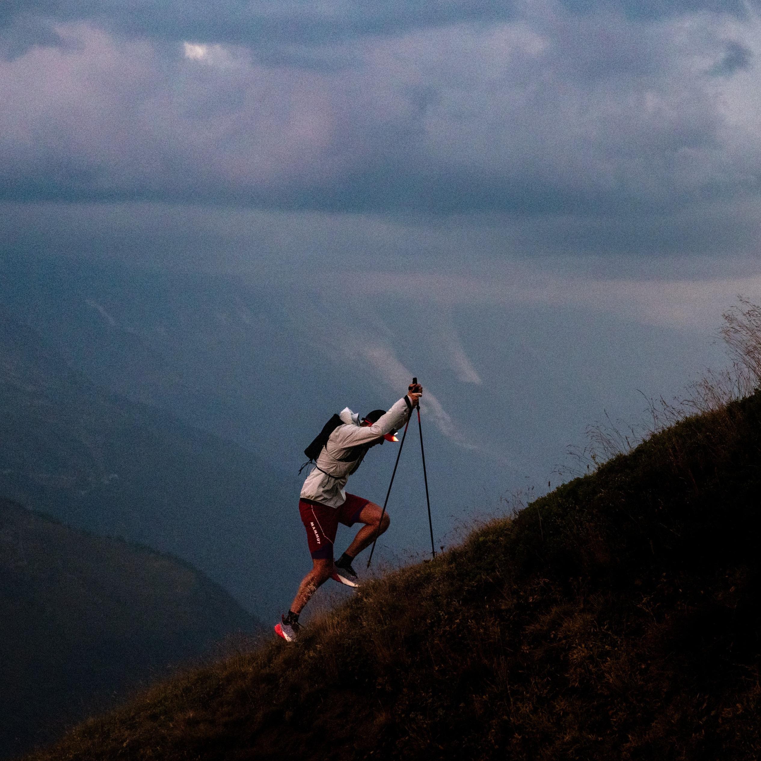 A hiker in Mammut gear ascends a mountain trail with trekking poles, under a cloudy sky, overlooking a vast alpine landscape.