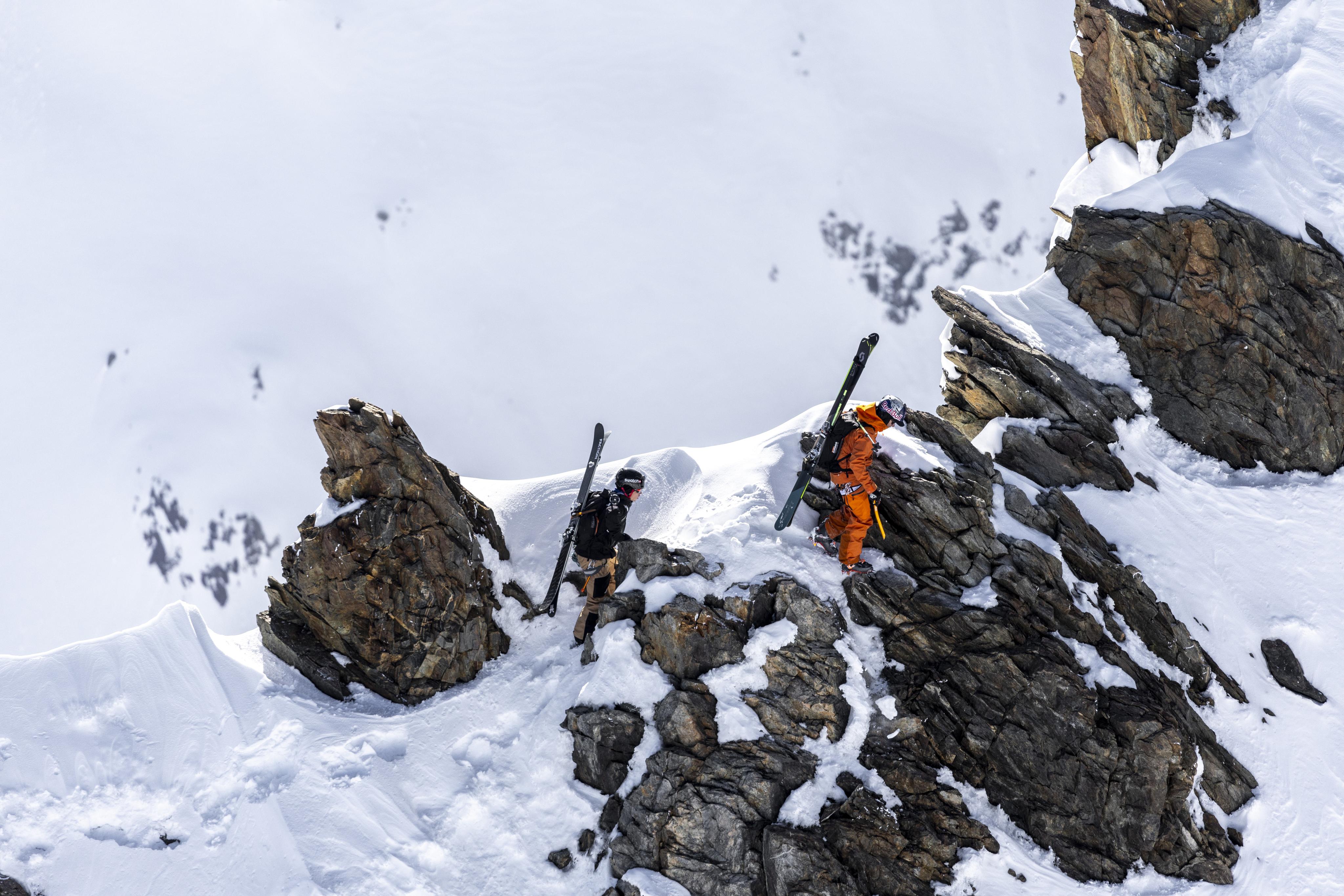 Two skiers in Mammut snow gear navigate a rugged, snowy mountain terrain, carrying Mammut skis over their shoulders.