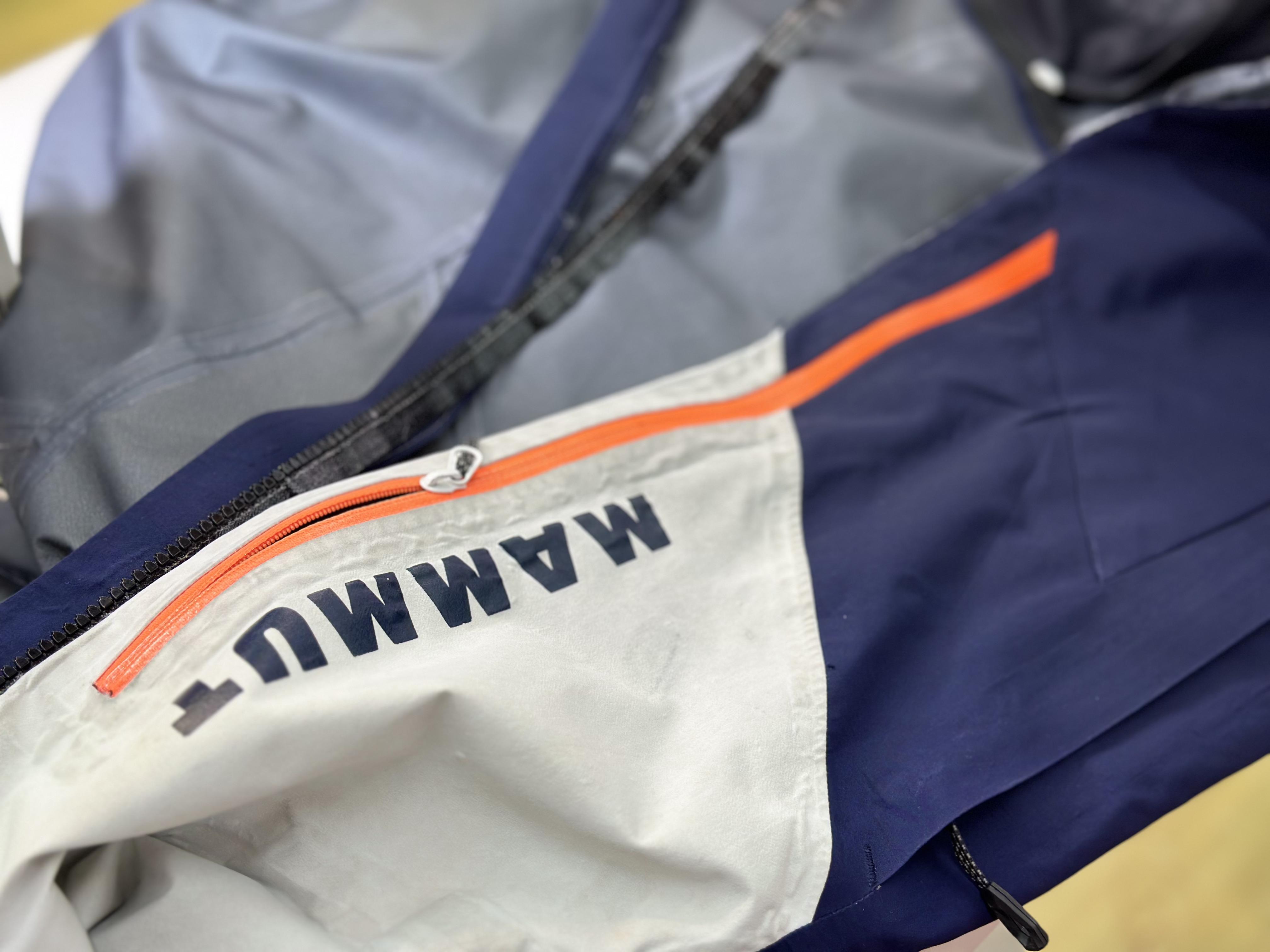 Close-up of a blue and gray Mammut jacket featuring an orange zipper and the word "Mammut" prominently displayed in black letters.